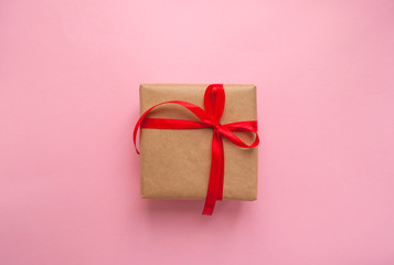 Gift box wrapped in brown colored craft paper and tied with red bow on pink background.