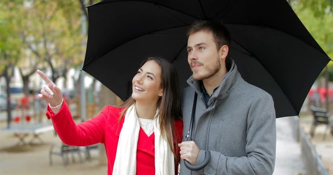 Front view of a happy couple walking towards camera and pointing away under the rain in winter
