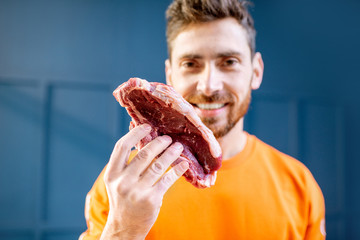Conceptual portrait of a man dressed in bright sweater with raw meat steak on the blue background