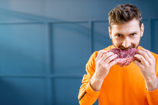Conceptual portrait of a man dressed in bright sweater biting raw meat steak on the blue background. Image with copy space