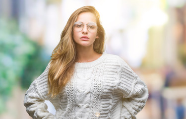 Young beautiful blonde woman wearing winter sweater and sunglasses over isolated background with serious expression on face. Simple and natural looking at the camera.