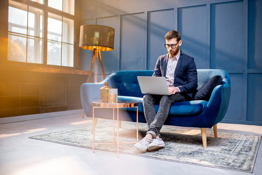 Portrait of an elegant businessman sitting with laptop on the couch at the luxury blue office interior
