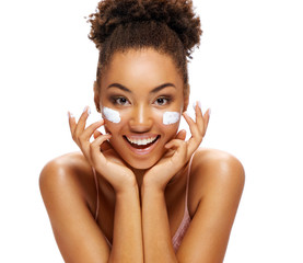 Happy young girl applying moisturizing cream on her face. Photo of smiling african american girl on white background. Skin care and beauty concept