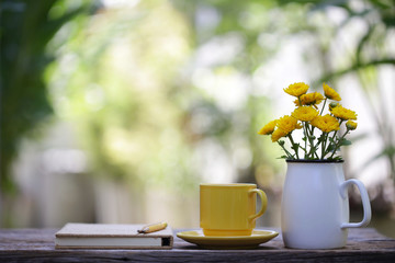 Yellow ceramic cup with flower jar and notebooks at on wooden table 