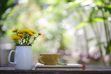 Yellow ceramic cup with flower jar and notebooks at on wooden table 