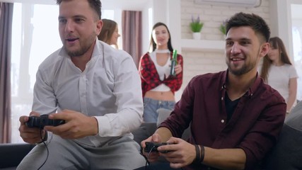 young gamers friends male play video games sitting on couch and funny girlfriends in background at home party