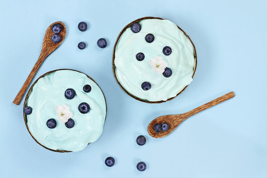 Blue Spirulina And Berry Smoothie Bowl, Fresh Fruit With Wooden Spoons Served In Coconut Bowls Over A Blue Background With White Flowers. Image Shot From Above / Overhead.