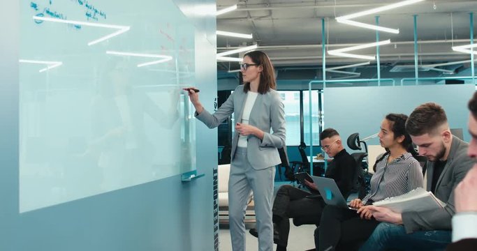 Attractive mid-30s female manager leading creative brainstorming meeting in modern office, presenting her ideas to the team on white board. 4K UHD