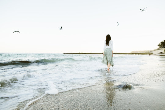 Marine female portrait. Attractive woman in green skirt walks along the shore before the sea while gulls fly over her