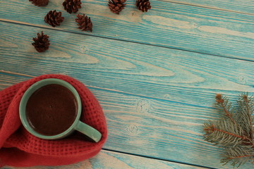 Coffee mug wrapped in a scarf, pine branch and cones on a blue background.	