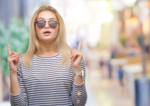 Young caucasian woman wearing sunglasses over isolated background amazed and surprised looking up and pointing with fingers and raised arms.