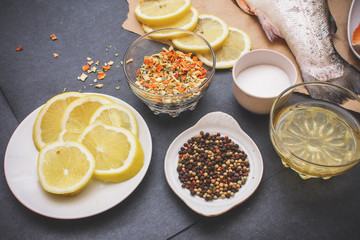 On dark gray paper a plate with lemon slices a plate with pepper with different color together with lemon salt and a glass of oil the tail of raw fish.