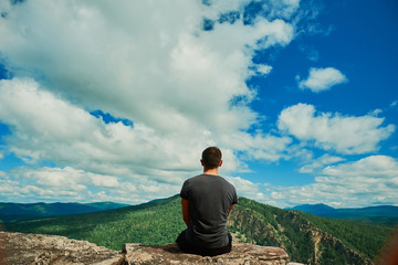 The guy sits back on the edge of the mountain. A man in gray t-shirt sits with his back overlooking...