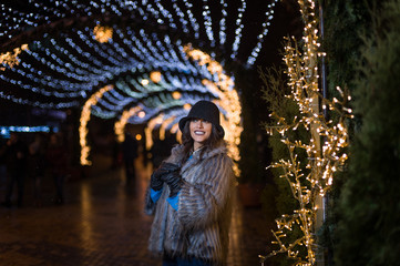 Pretty dark haired girl wearing a fur coat, blue jeans, blue top and a black hat, smiling, posing with snowflakes Christmas lights outdoor at night time.