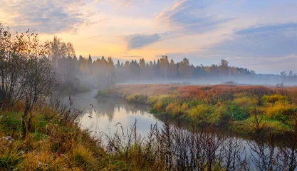 Papier Peint photo Automne Foggy autumn landscape with small forest river.Autumnal hazy early morning.Twilight.Calm stream of river flowing between the woods and meadows covered by fog.Colorful clouds in sky at sunrise.