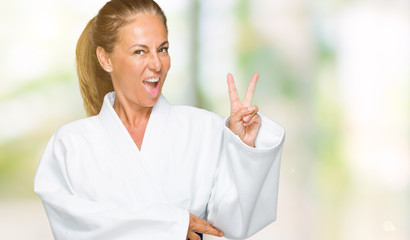 Middle age adult woman wearing karate kimono uniform over isolated background smiling with happy face winking at the camera doing victory sign. Number two.