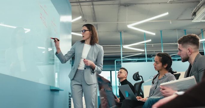 Attractive mid-30s female manager leading creative brainstorming meeting in modern office, presenting her ideas to the team on white board. 4K UHD