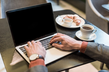 cropped view of businessman sitting at table with coffee and croissant while typing on laptop with blank screen in restaurant