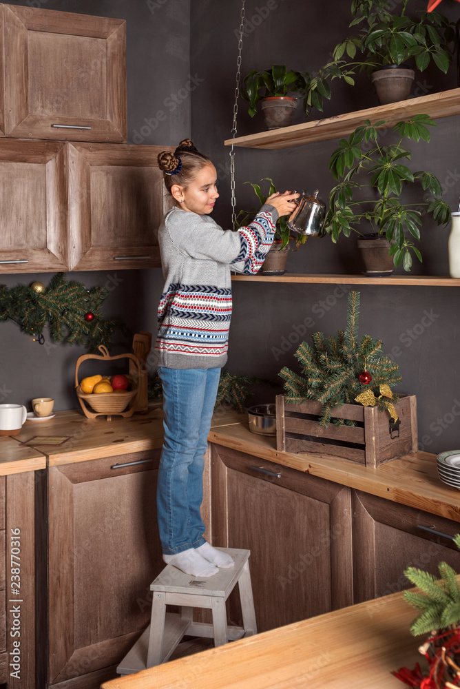 Wall mural teenager girl in a sweater and jeans stands on a stool and watering flowers on the upper shelves of 