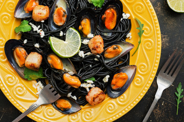 Closeup of black spaghetti with mussels, scallops, lime, mozzarella cheese and spices on a yellow plate. Top view