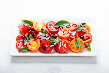 Light dietary salad with organic tomatoes, balsamic vinegar, olive oil, salt and basil leaves in a rectangular dish on a white wooden board. The concept of dietary nutrition