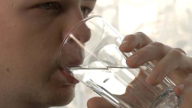 Close up shot of a young man drinking transparent soda from a glass in front of the window
