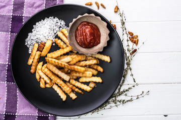 Plate of homemade french potatoes with ketchup and sea salt on plaid napkin and white wooden background, top view