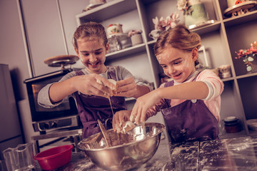 Cute happy sisters adding eggs to the dough
