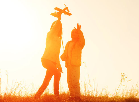 Two little sisters holding hands and playing with small airplane toy outdoors. Silhouette of two sisters walking on the meadow at sunset