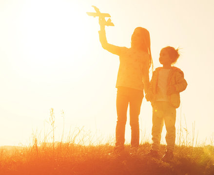 Two little sisters holding hands and playing with small airplane toy outdoors. Silhouette of two sisters walking on the meadow at sunset