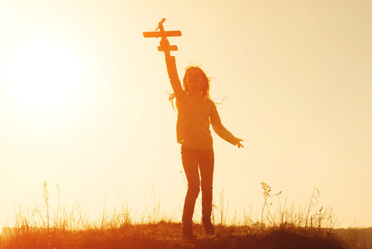 Little Girl's Silhouette Holding Small Toy Airplane And Smiling. Cheerful Girl Standing The Meadow At Sunset Time