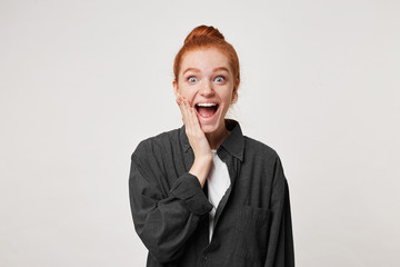 Surprised positive female with blue eyes popped out, palm touching cheek, opens mouth widely in surprisement as hears news from interlocutor, straes at camera, expresses shock and disbelief