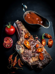 Fototapeta na wymiar Grilled hot ribeye beef steak on bone with spicy basting sauce and tomatoes on dark cooking background