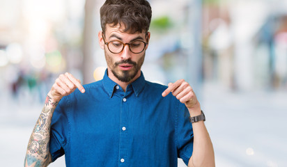 Young handsome man wearing glasses over isolated background Pointing down with fingers showing advertisement, surprised face and open mouth