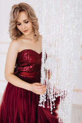 Fototapeta premium Adorable woman in red burgundi dress poses in a bright luxury room with large chandelier