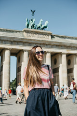 Portrait of a young beautiful positive smiling stylish tourist girl in Berlin in Germany. Brandenburg Gate and unrecognizable blurred people in the background.