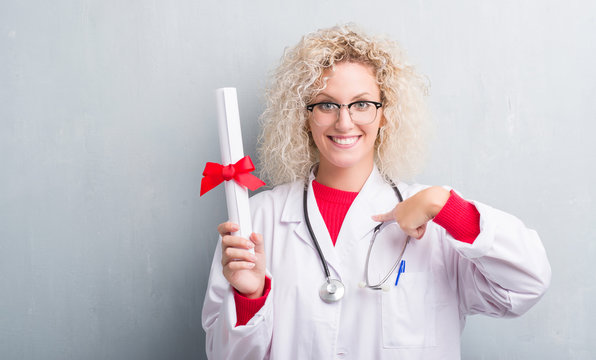 Young blonde doctor woman over grunge grey wall holding degree with surprise face pointing finger to himself