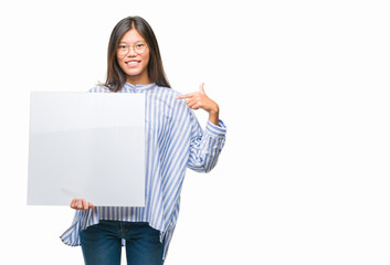 Young asian woman over isolated background holding blank banner with surprise face pointing finger to himself