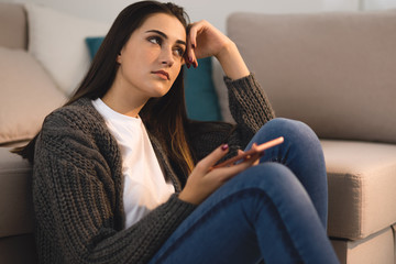 Depressed woman sitting at home and looking away while waiting for a call from her boyfriend