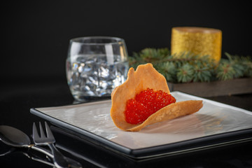 Studio shot of red caviar on a tartlet on a square black white plate.  Nearby are  a candle with Christmas tree branches and tableware