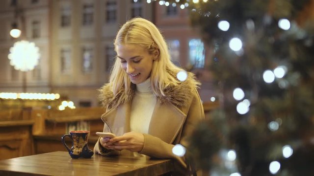Woman using smartphone on christmas market. Girl enjoying winter holiday season. Concept of  social networking, communicating, using app. Blurred Christmas lights on background.
