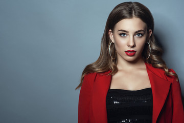 Portrait of beautiful lady with perfect make up and shiny hair tucked behind her ears wearing black sequin top and bright red jacket. Luxurious hoop earrings. Isolated on blue background. Text space © obrik