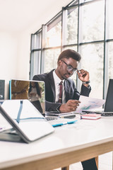 Successful young African executive sitting at his desk in an office smiling and working on a laptop