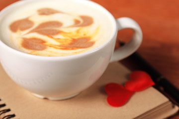 Valentine's day background. A white cappuccino Cup with a heart-shaped latte art stands on an old notebook with sheets of paper, on a wooden table with a handle and soft silk hearts close-up
