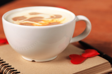 Valentine's day background. A white cappuccino Cup with a heart-shaped latte art stands on an old notebook with sheets of paper, on a wooden table with a handle and soft silk hearts close-up