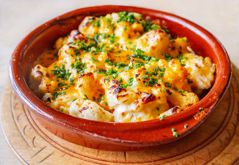 Baked cauliflower cheese made with Red Leicester served in a rustic red ceramic round dish. Cooked with a gratin top and sprinkled with parsley. The dish is on a wooden serving board