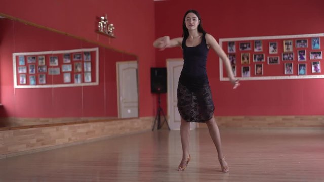 Fit slim young brunette with long  black straight hair in tight black dress with long legs showing moves of salsa/bachata/chacha. Teaching dancing. Dancing to the right/left. Red walls background
