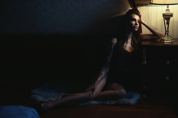 Gorgeous sad tattooed lady in black lingerie sitting on the floor at the hotel leaning on the bedside table and looking aside. Loneliness concept. Text space