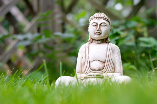 Small white Buddha statue in a meditation pose with green grass foreground and on natural bright blurred background. Religious symbol of buddhism