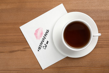 Paper napkin with phone number and lipstick mark under tea cup on wooden table, top view